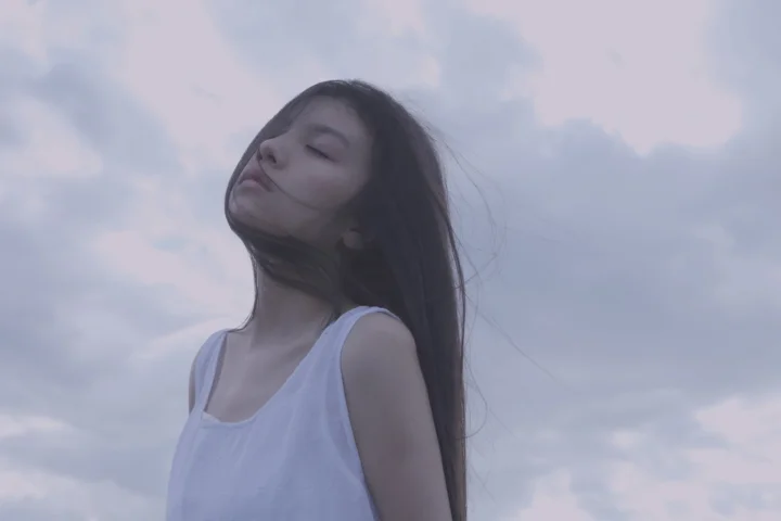 一名长发女子闭眼站在云天下，微风吹动她的头发。