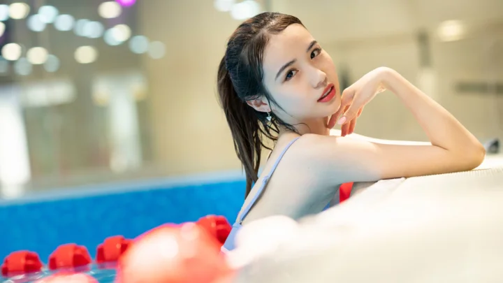 한 여성이 수영장 가장자리에 기대어 있고 배경에는 부드럽게 흐려진 불빛이 보입니다.