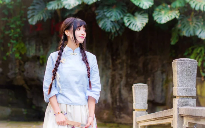 一名身穿浅色传统服饰、留着双辫的女性站在绿叶和石栏旁。