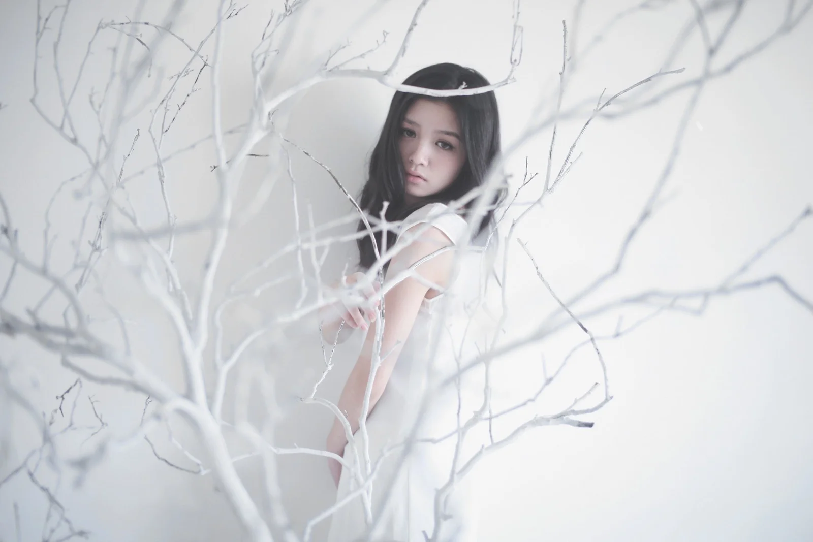 一名身穿白色服装的成年女子站在白色枝条之间，画面以浅色调为主。
