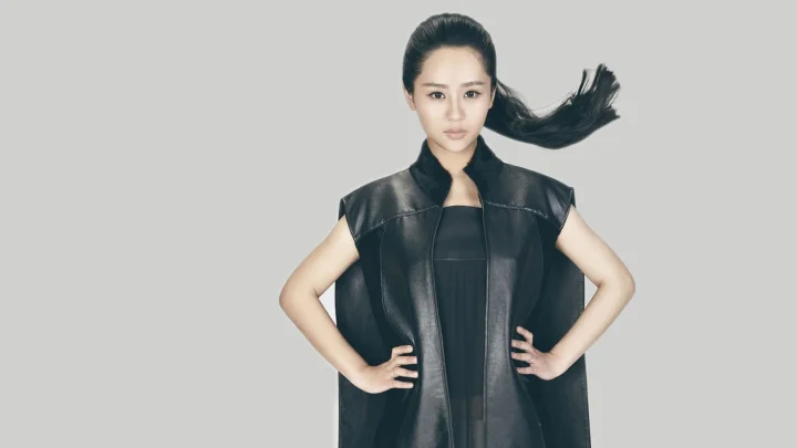 一名成年女性身穿黑色无袖外套站在灰色背景前，双手叉腰，长发向右侧飘起。