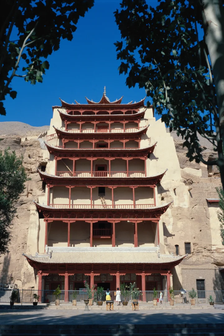 一座红色多层古建筑坐落在岩壁前，周围被树木环绕。