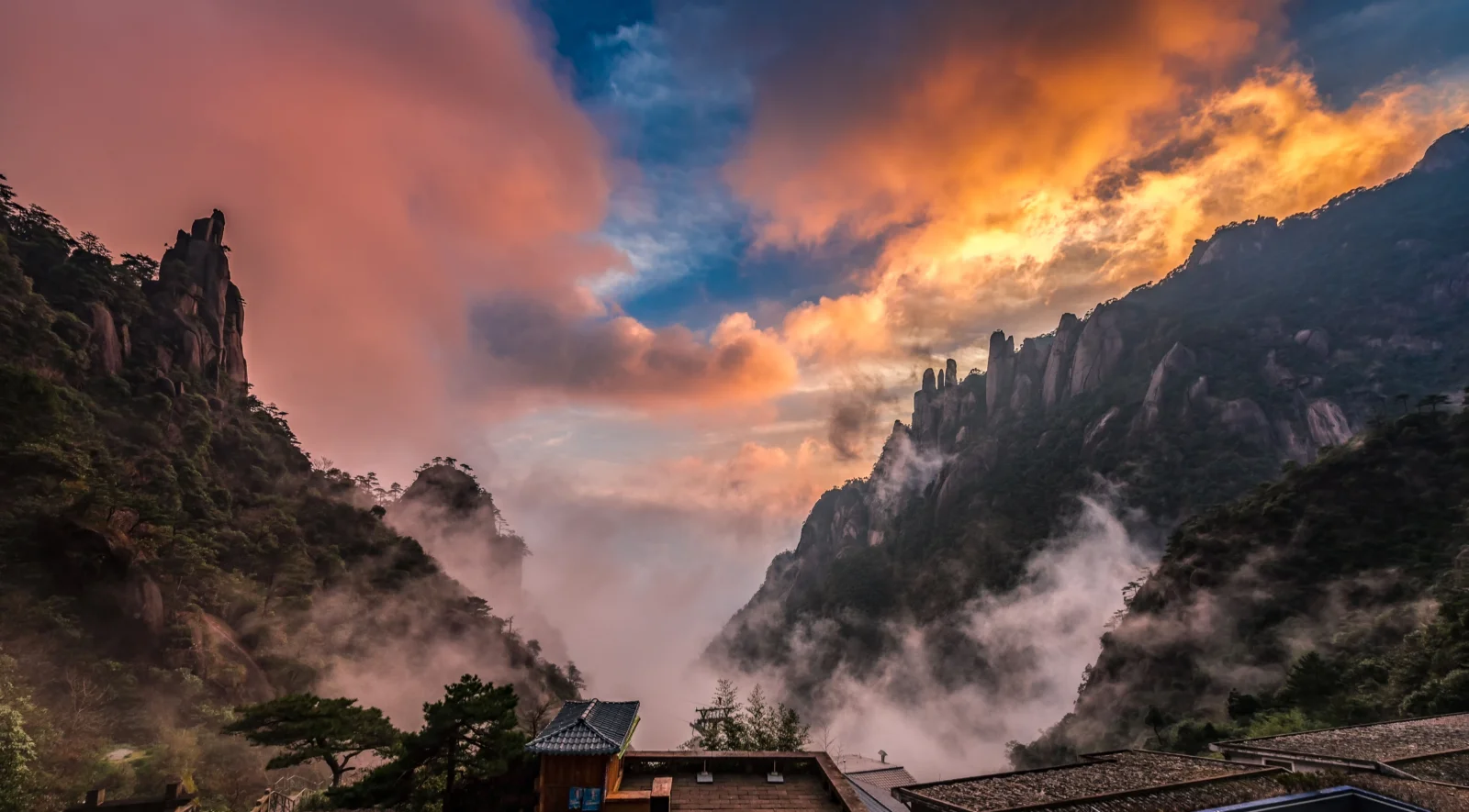 群山、缭绕的雾气和晚霞映照的云层构成壮丽的自然景观，前景可见少量屋顶。