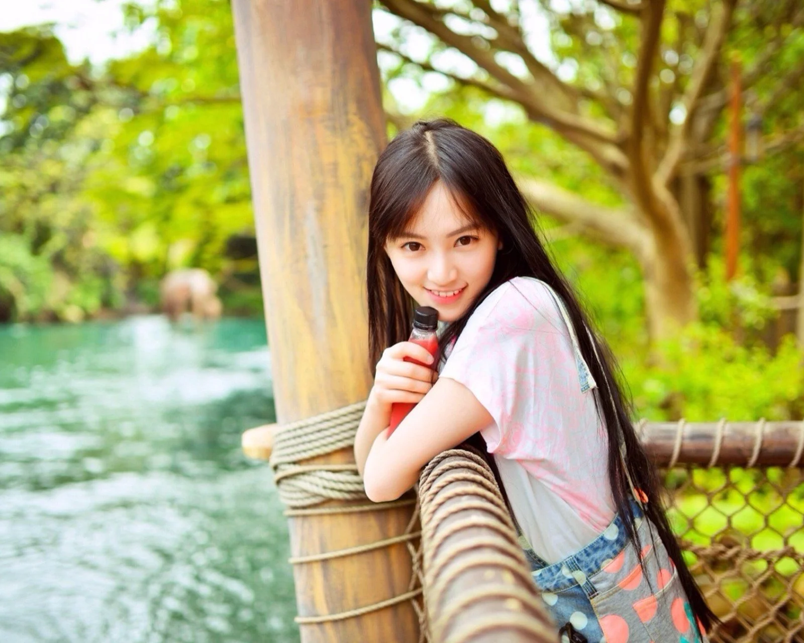 一名女子倚在河边的竹制栏杆旁，周围是绿树和流动的水面。