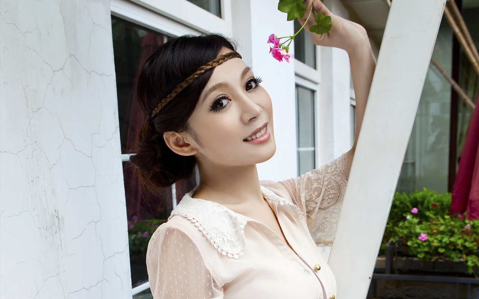 一名成年女子站在窗边，手持一枝粉色小花微笑面对镜头。