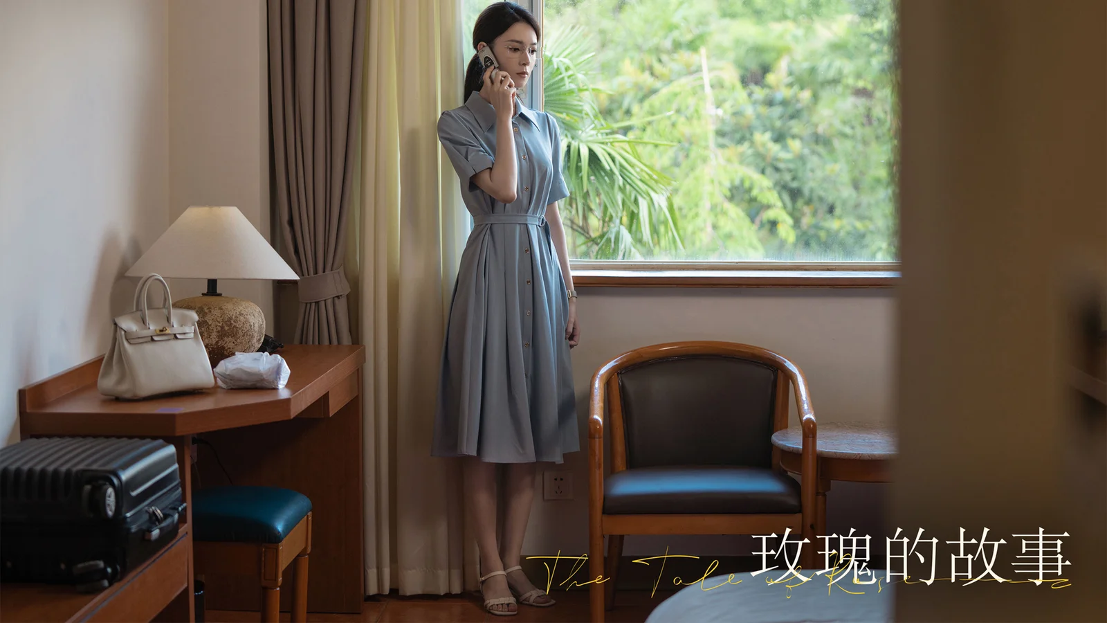 一名成年女子站在室内窗边，身旁可见椅子、桌子和行李箱。