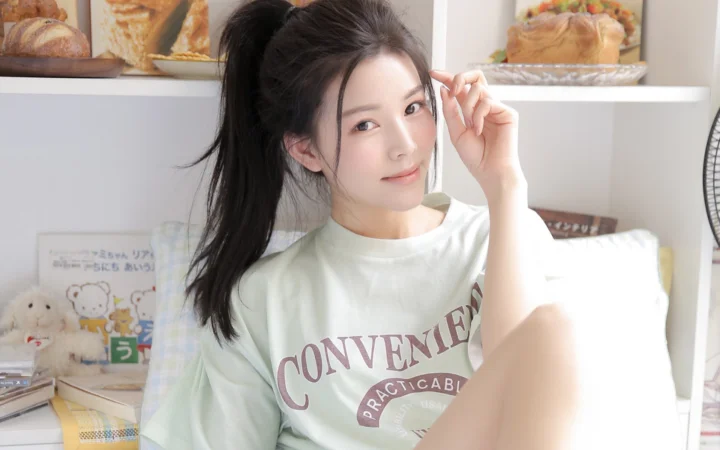 一名扎着马尾的女性穿着白色印花T恤坐在室内，身后是摆放着面包和玩偶的架子。