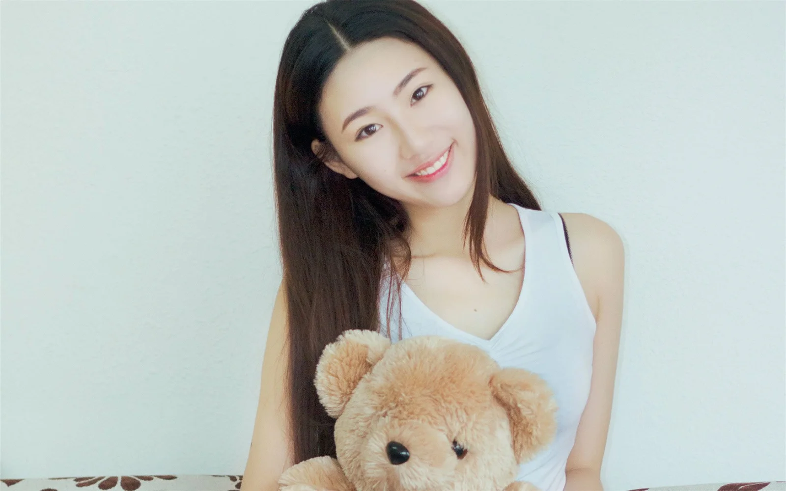 긴 머리의 여성이 밝은 색 벽을 배경으로 갈색 테디베어를 안고 미소 짓고 있다.