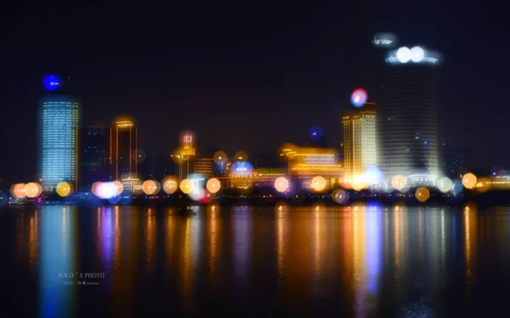 夜色中的城市建筑灯光倒映在平静水面上，呈现出柔和的散景效果。