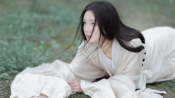 一名长发女子穿着白色服装，俯身躺在草地上。