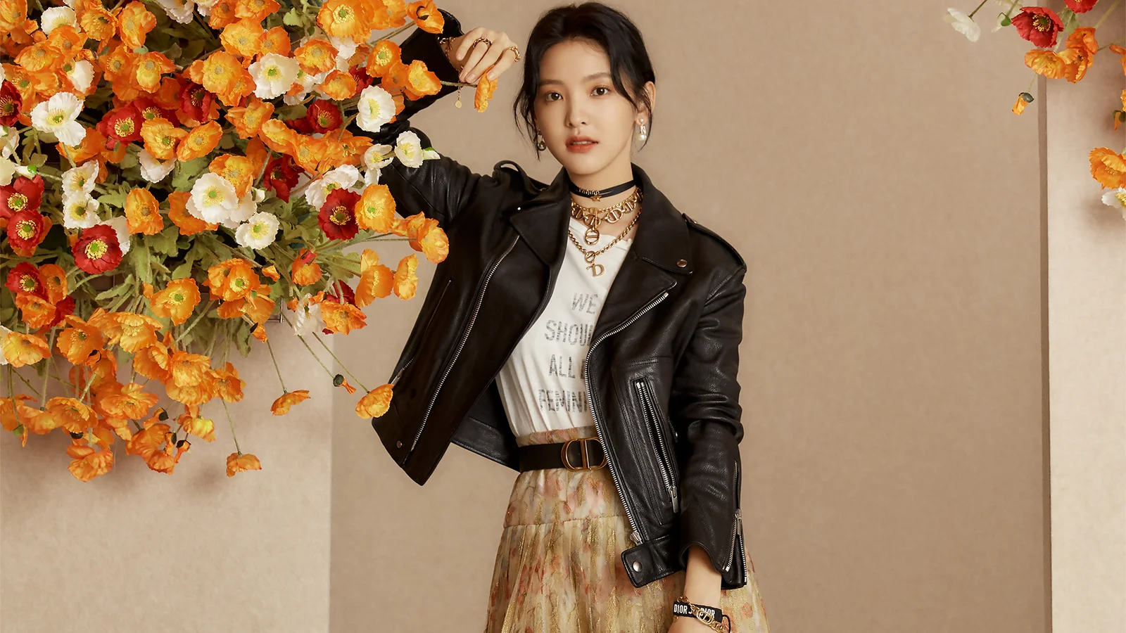 검은색 가죽 재킷을 입은 성인 여성이 주황색과 흰색 꽃을 배경으로 서 있다.