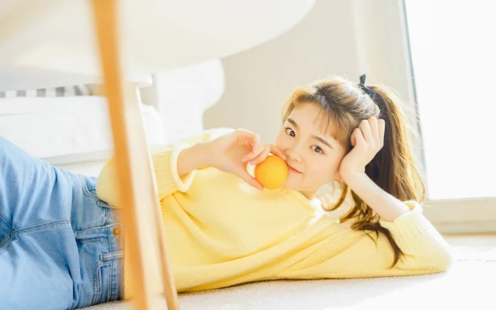 노란색 상의를 입은 여성이 밝은 실내에서 오렌지를 손에 들고 누워 있습니다.