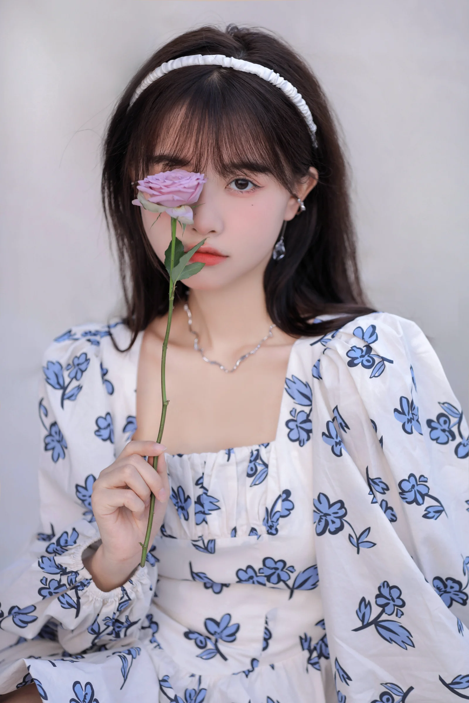 一名戴白色发带的女性身穿蓝色花卉图案连衣裙，手持一枝玫瑰面对镜头。
