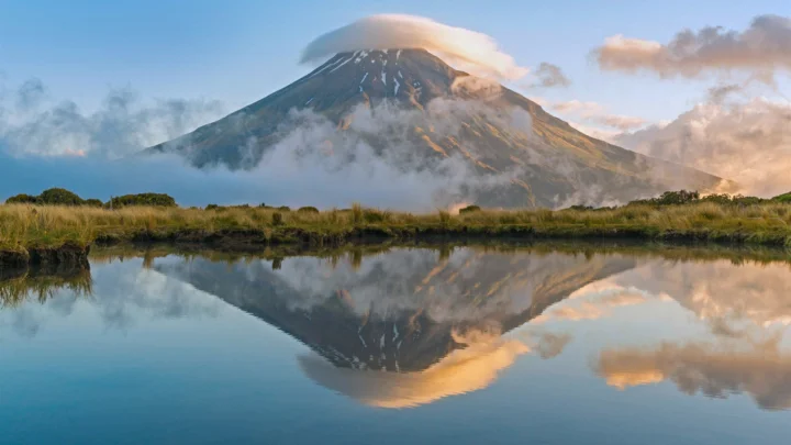 一座云雾缭绕的山峰倒映在平静的湖面上，前景是草地与水岸。