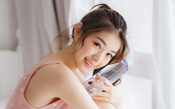 一位身穿粉色上衣的成年女性在室内微笑并手持透明瓶子。