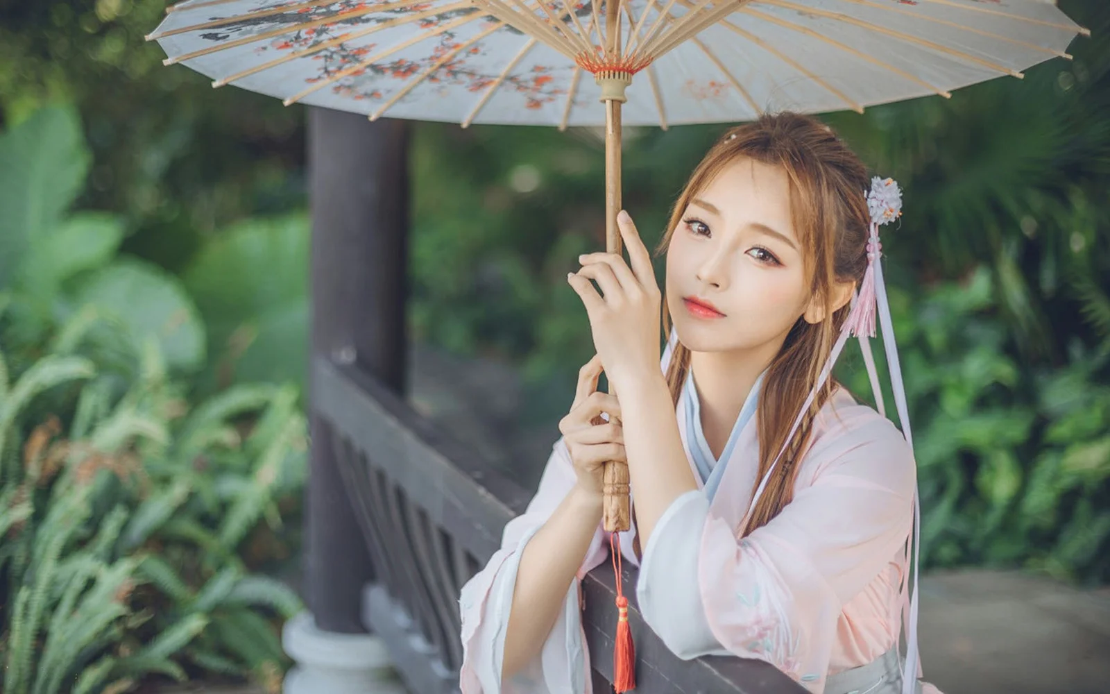 푸른 식물에 둘러싸인 야외에서 한 여성이 연한 색의 양산을 들고 있다.