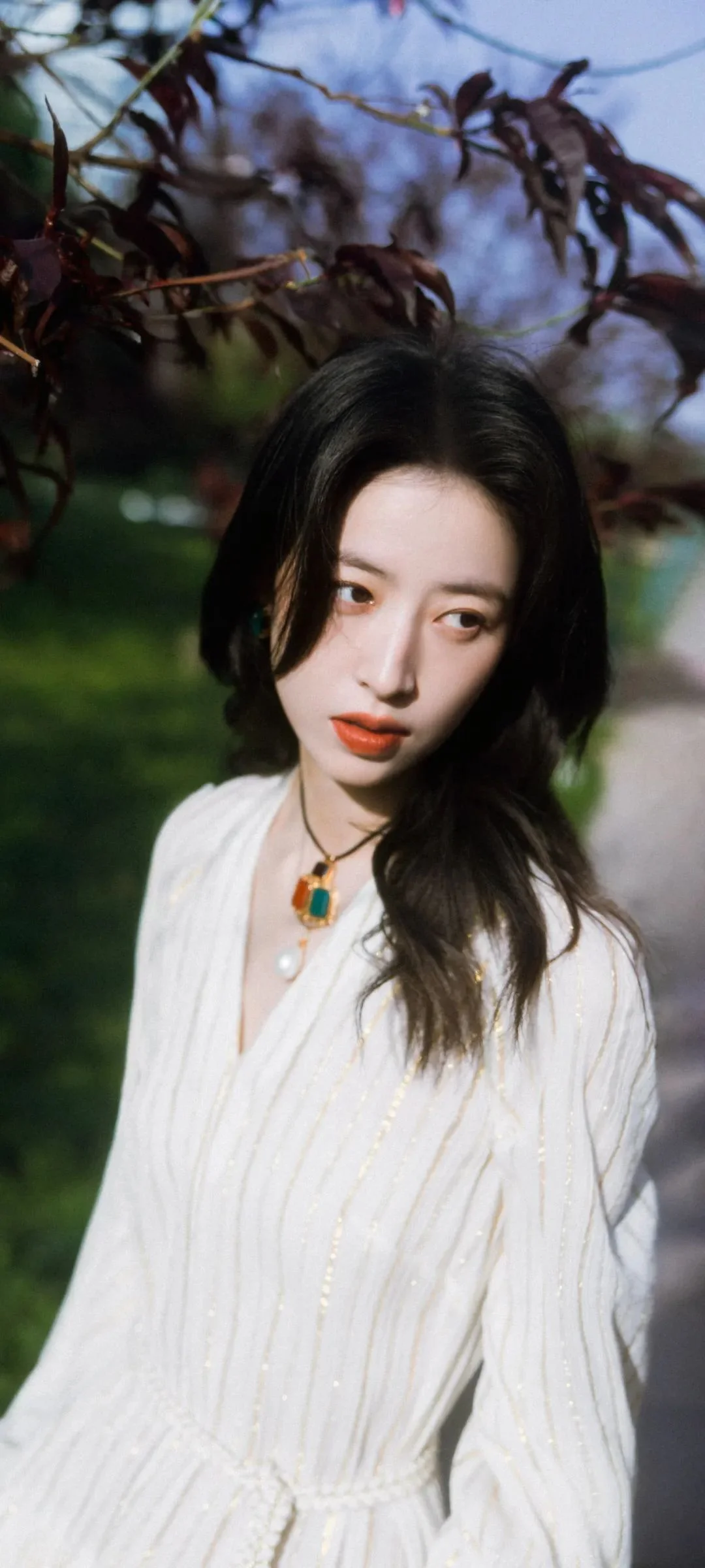 흰색 원피스를 입은 여성이 나뭇가지와 초록 식물로 둘러싸인 야외에 서 있습니다.