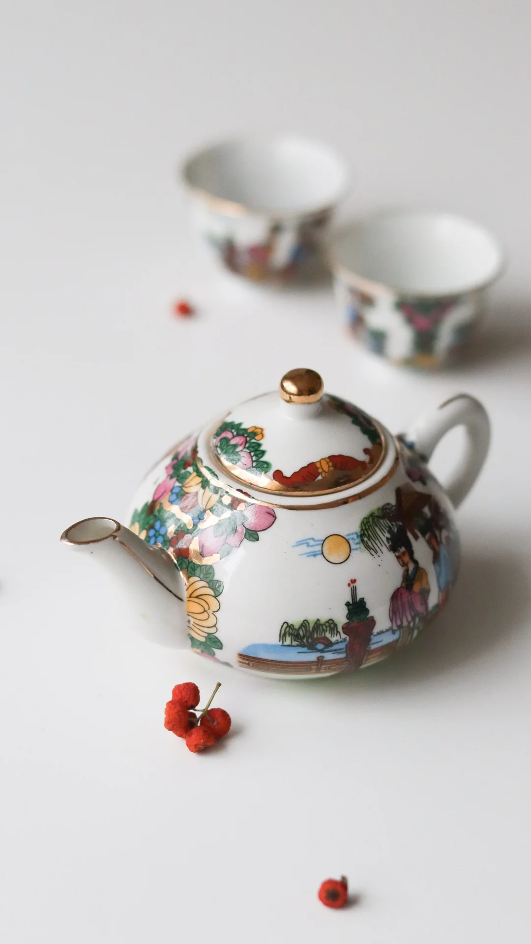 白色桌面上摆放着一只彩绘花卉茶壶、两只茶杯和几颗红色果实。