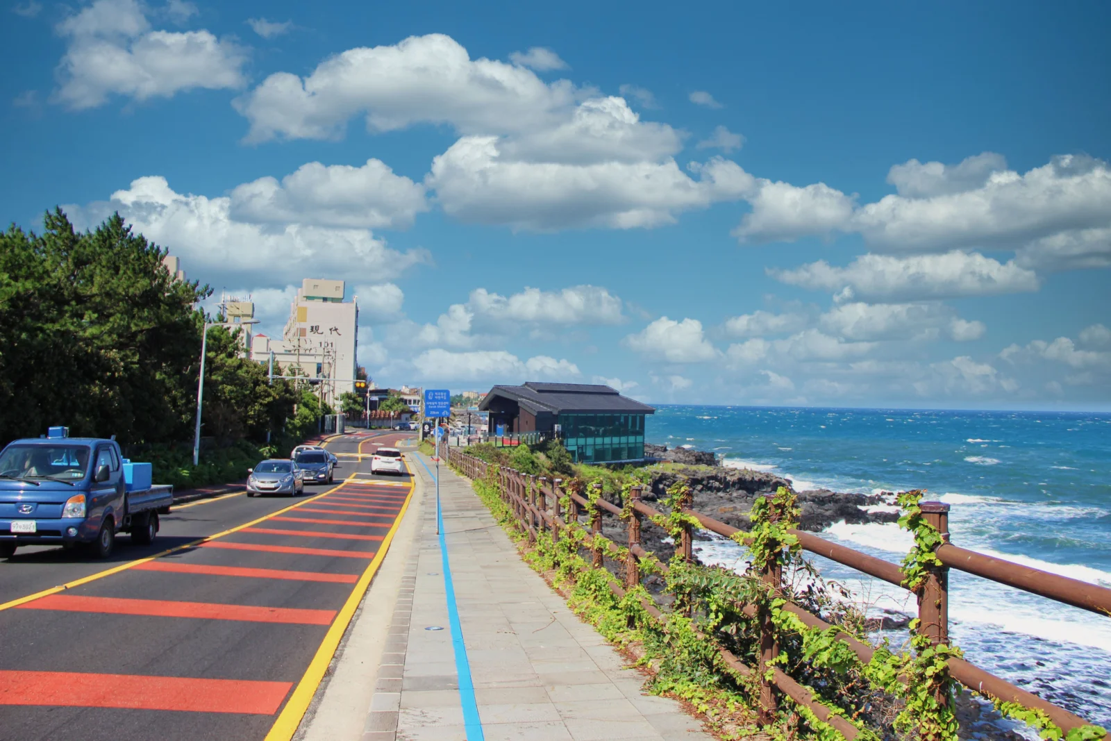 白い雲が浮かぶ青空の下、青い海沿いに海岸道路が伸び、木々や建物、海辺の遊歩道が見える。
