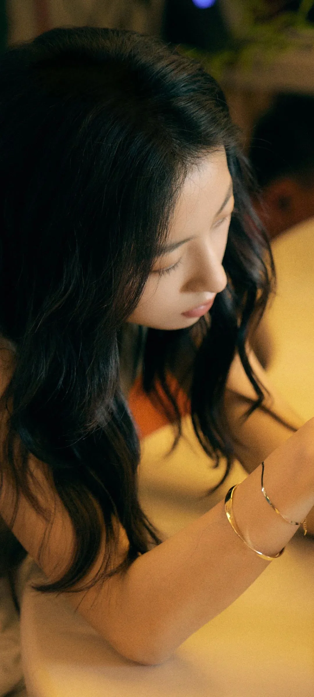 긴 머리의 여성이 따뜻한 빛 속에서 노란색 상의를 입고 아래를 바라보고 있다.
