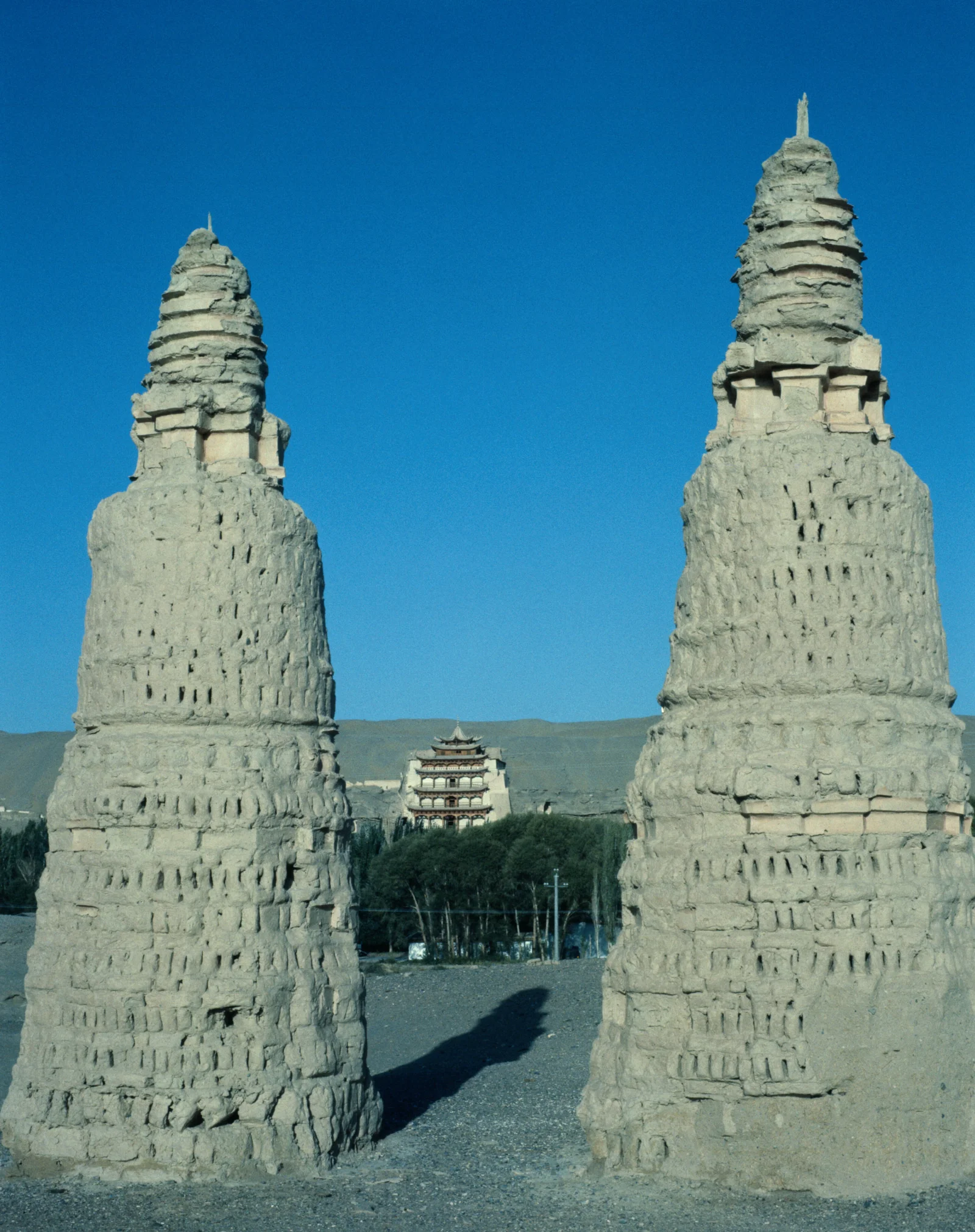 两座高大的石塔矗立在前景，中间远处可见一座多层古建筑，背景是蓝天与山丘。