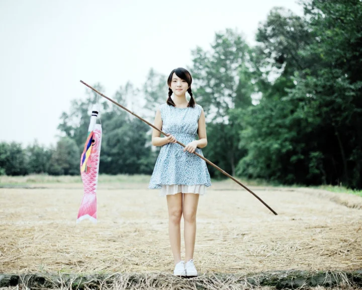 一名女子站在铺满稻草的田野中，手持一根长杆，身后是树木。