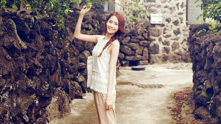 一名成年女子站在石墙环绕的小径旁，面带微笑并抬手触碰上方的枝叶。