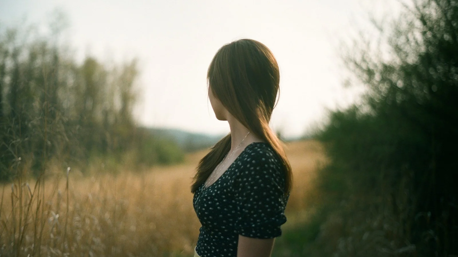 한 여성이 나무로 둘러싸인 황금빛 들판에 서서 먼 곳을 바라보고 있습니다.