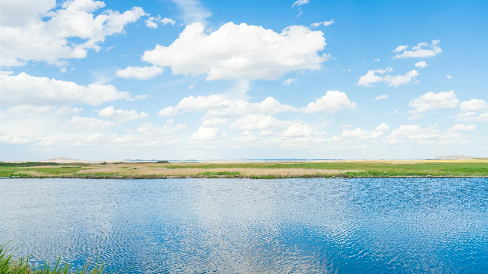 画面展现了蓝天下的湖面、云朵和远处的绿色草地。