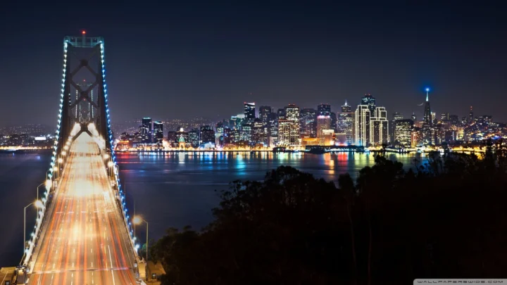夜色中，一座大桥通向灯火璀璨的城市天际线，水面倒映着绚丽灯光。