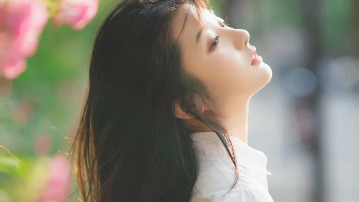 야외에서 꽃과 초록빛에 둘러싸인 성인 여성이 옆모습으로 위를 바라보고 있다.