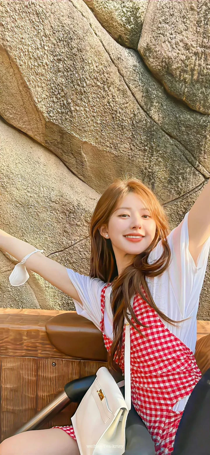 긴 머리의 여성이 바위 배경 앞에 앉아 빨간색과 흰색 체크무늬 원피스와 흰색 상의를 입고 있다.