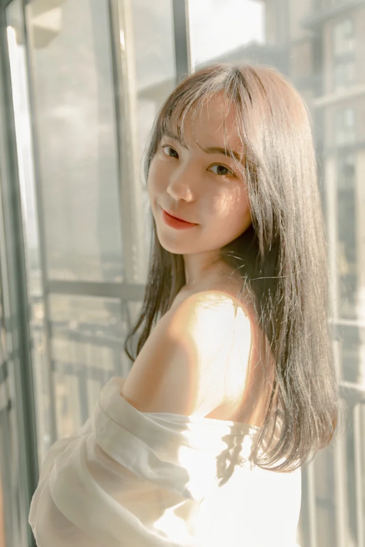 一位长发女性站在阳光照射的窗边。