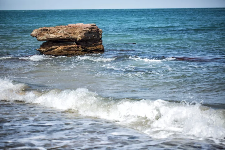 海浪拍打着浅色海岸，中央是一块突出的岩石，远处延伸着蓝绿色海面。