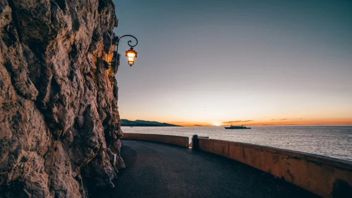 海边悬崖旁的道路延伸向宁静海面，远处夕阳映照着橙色天空。