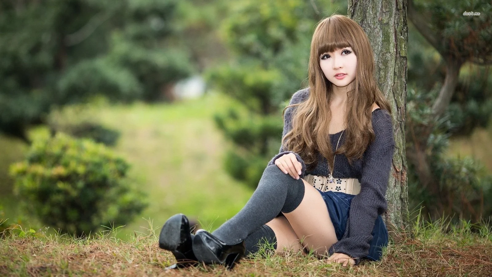 성인 여성이 나무 옆 잔디에 앉아 있으며 배경에는 부드러운 녹색 식물이 보입니다.