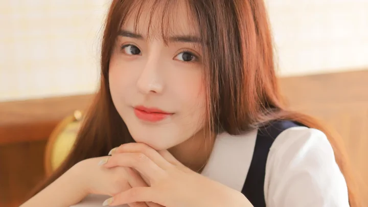 一名棕色长发女性靠在桌前，双手交叠并面向镜头微笑。