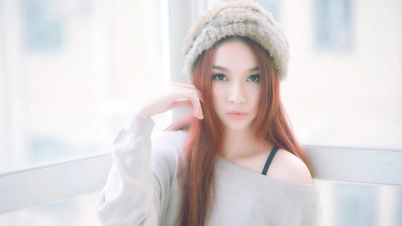 ニット帽をかぶった赤毛の女性が、明るい窓辺で淡い色のゆったりしたトップスを着て座っています。