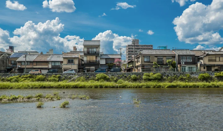 河流前方是一排日本传统民居，背后是城市建筑与蓝天白云。