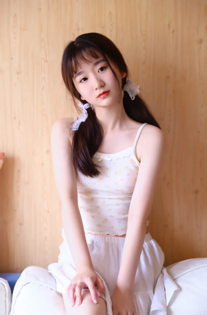 一名长发女性坐在室内木质背景前，身穿浅色吊带上衣和白色裙子。