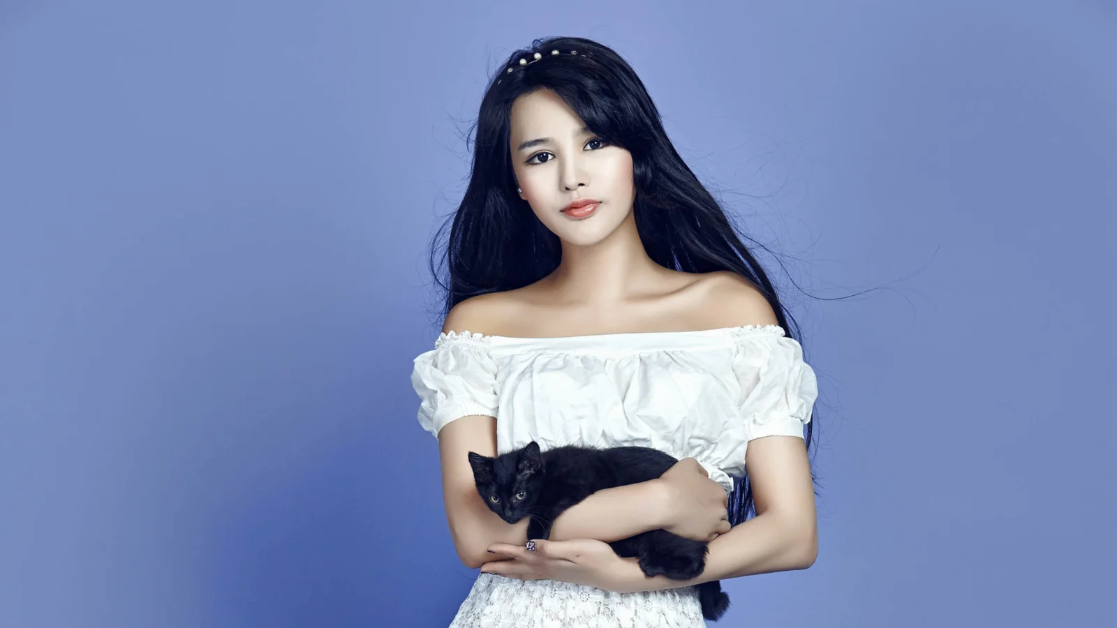 一名长发女性穿着白色露肩上衣，在蓝色背景前抱着一只黑猫。