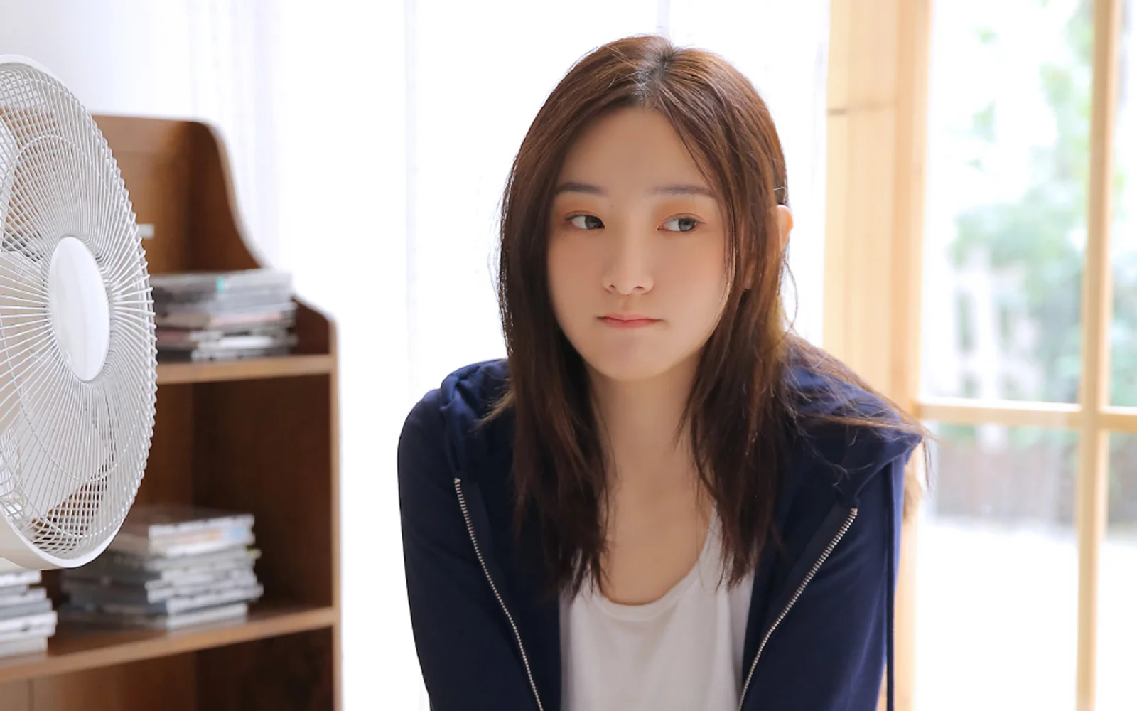 明るい室内で、扇風機と本棚のそばに成人女性が座っています。