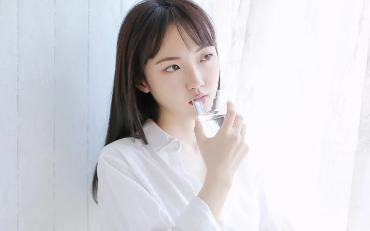 一名身穿白衬衫的成年女性在明亮窗边手持玻璃杯饮水。