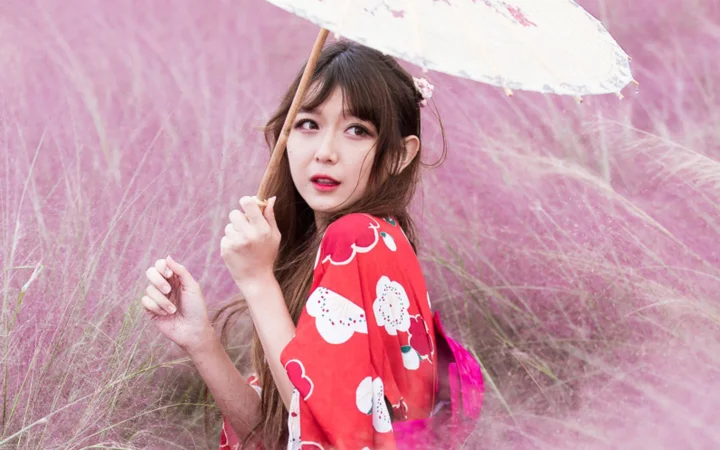 一名女子撑着白色伞站在粉色草丛中，身穿红色花纹服装。