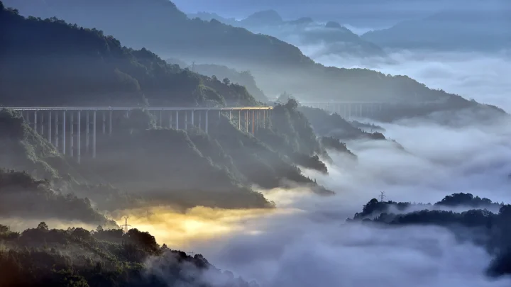 群山被云雾笼罩，一座高架桥横跨山谷，远处透出柔和的晨光。