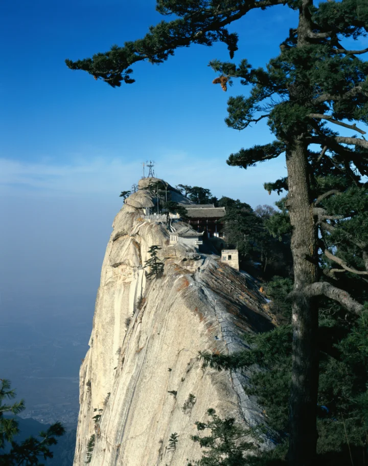 陡峭的岩峰被苍松环绕，峰顶可见一座建筑，远处笼罩在蓝色雾霭中。