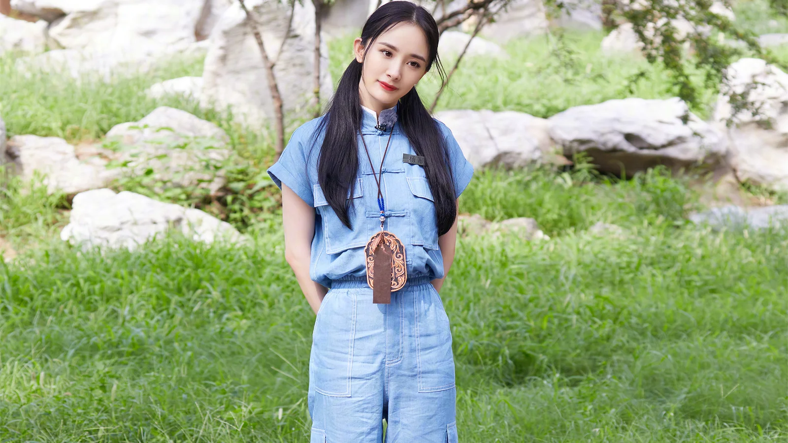 青い服を着た女性が、緑の草と岩に囲まれた屋外に立っている。
