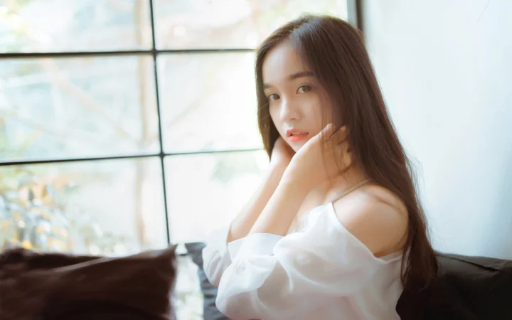 긴 머리의 여성이 창가에 앉아 흰색 오프숄더 상의를 입고 부드러운 햇빛을 받고 있습니다.