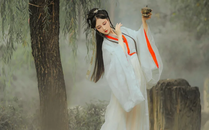 흰색 전통 의상을 입은 여성이 버드나무 가지와 나무 그루터기 사이의 야외 안개 낀 풍경에 서 있다.
