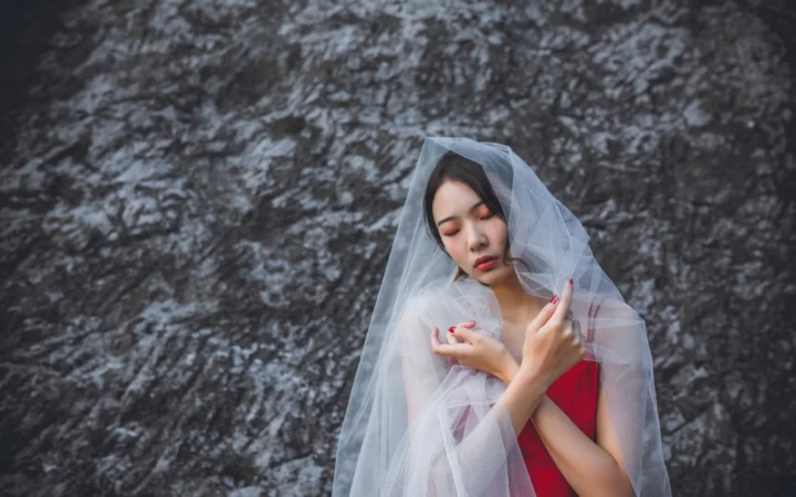 一名成年女性闭着眼睛，身披半透明薄纱并穿着红色服装，站在岩石墙前。